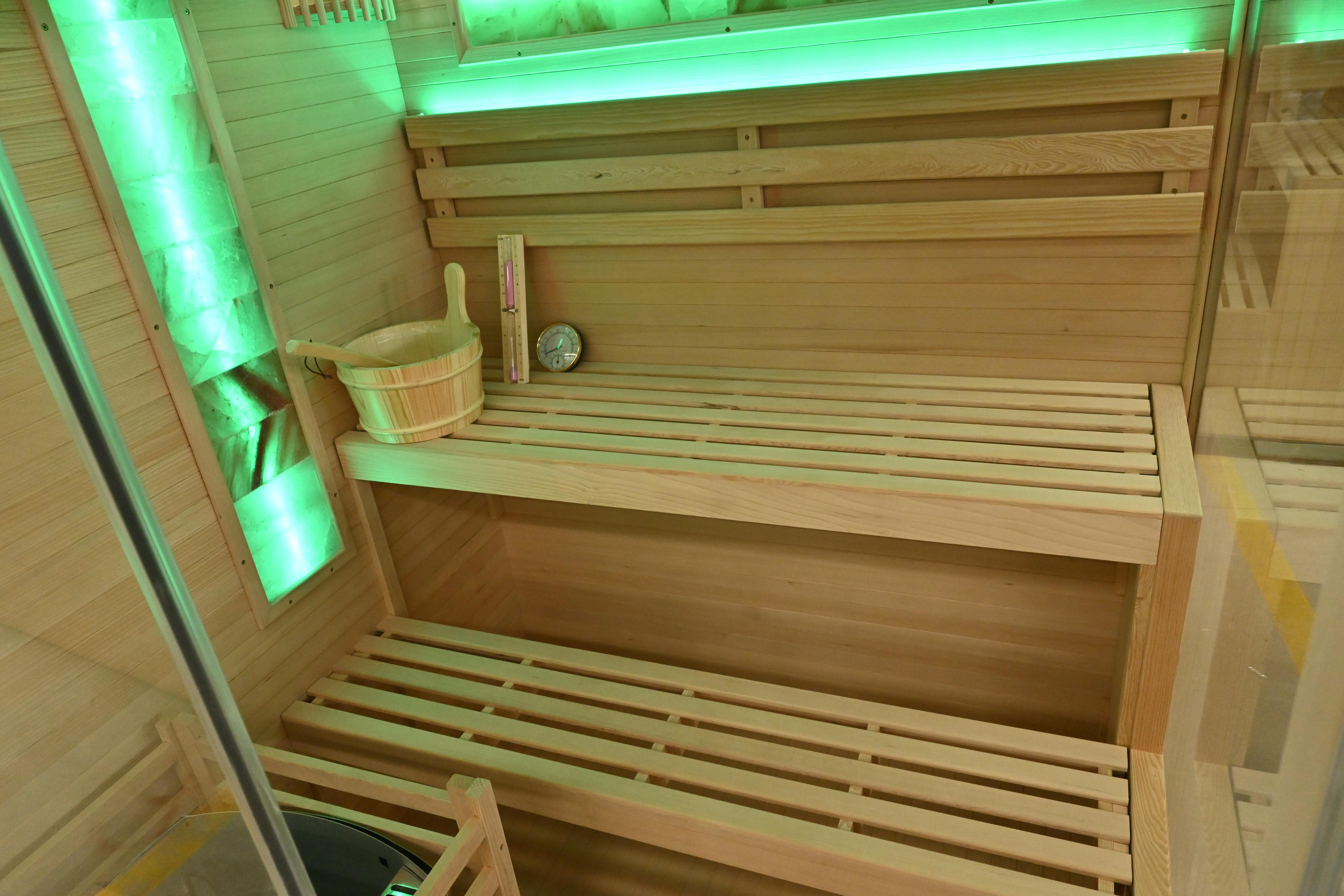 Innenraum einer Sauna mit Holzbänken, einem Eimer und einem Thermometer. Grüne Beleuchtung.