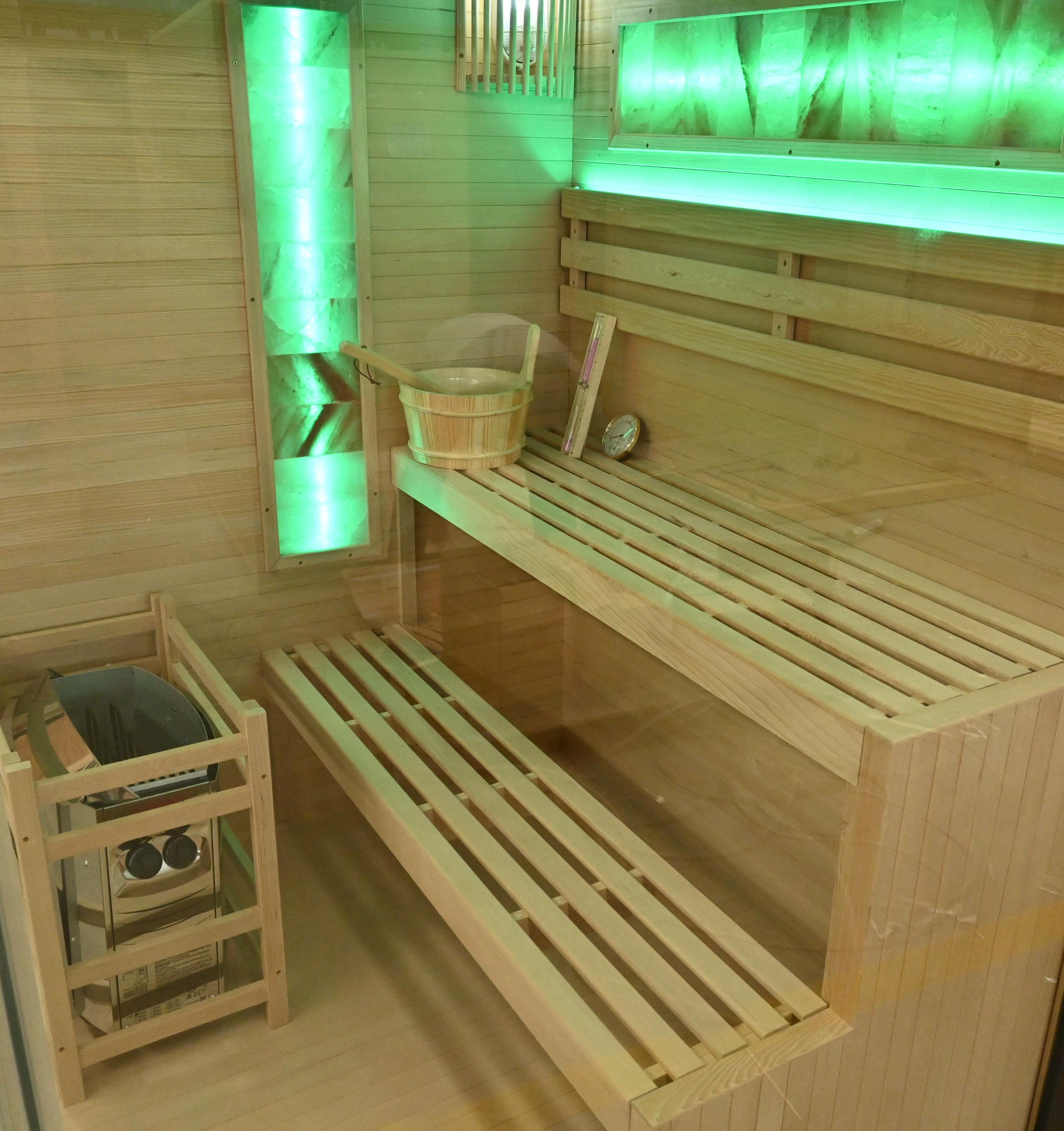 Innenraum einer Sauna mit Holzbänken, einem Heizgerät, einem Eimer und einem Thermometer. Grüne Beleuchtung verstärkt die Szene.