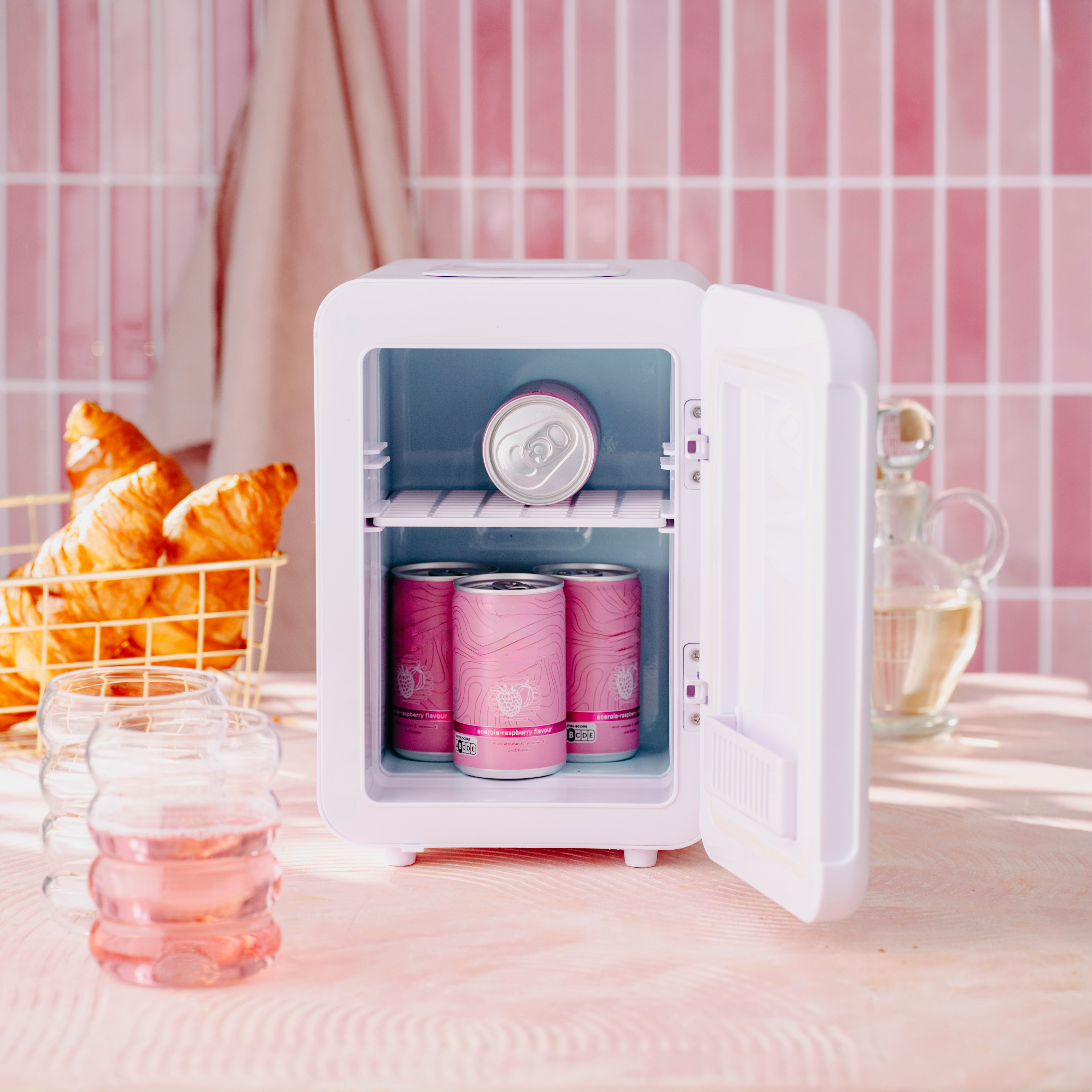 Egy fehér mini hűtőszekrény nyitott ajtóval. Belül rózsaszín dobozok vannak. Croissantok egy arany kosárban, poharak és rózsaszín csempés háttér.