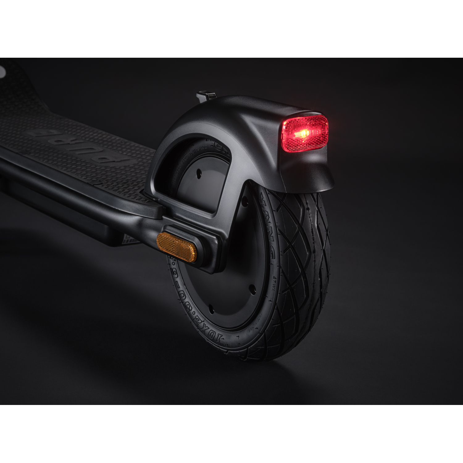 Gros plan d'une trottinette électrique noire. La roue arrière et un feu rouge ressortent sur fond sombre. Un réflecteur jaune est visible.