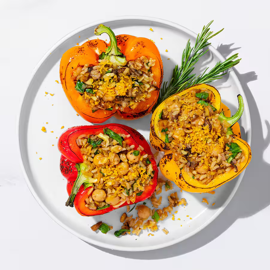 Három töltött paprika egy fehér tányéron. Egy piros, egy narancs és egy sárga. Rizskeverékkel és csicseriborsóval vannak töltve.