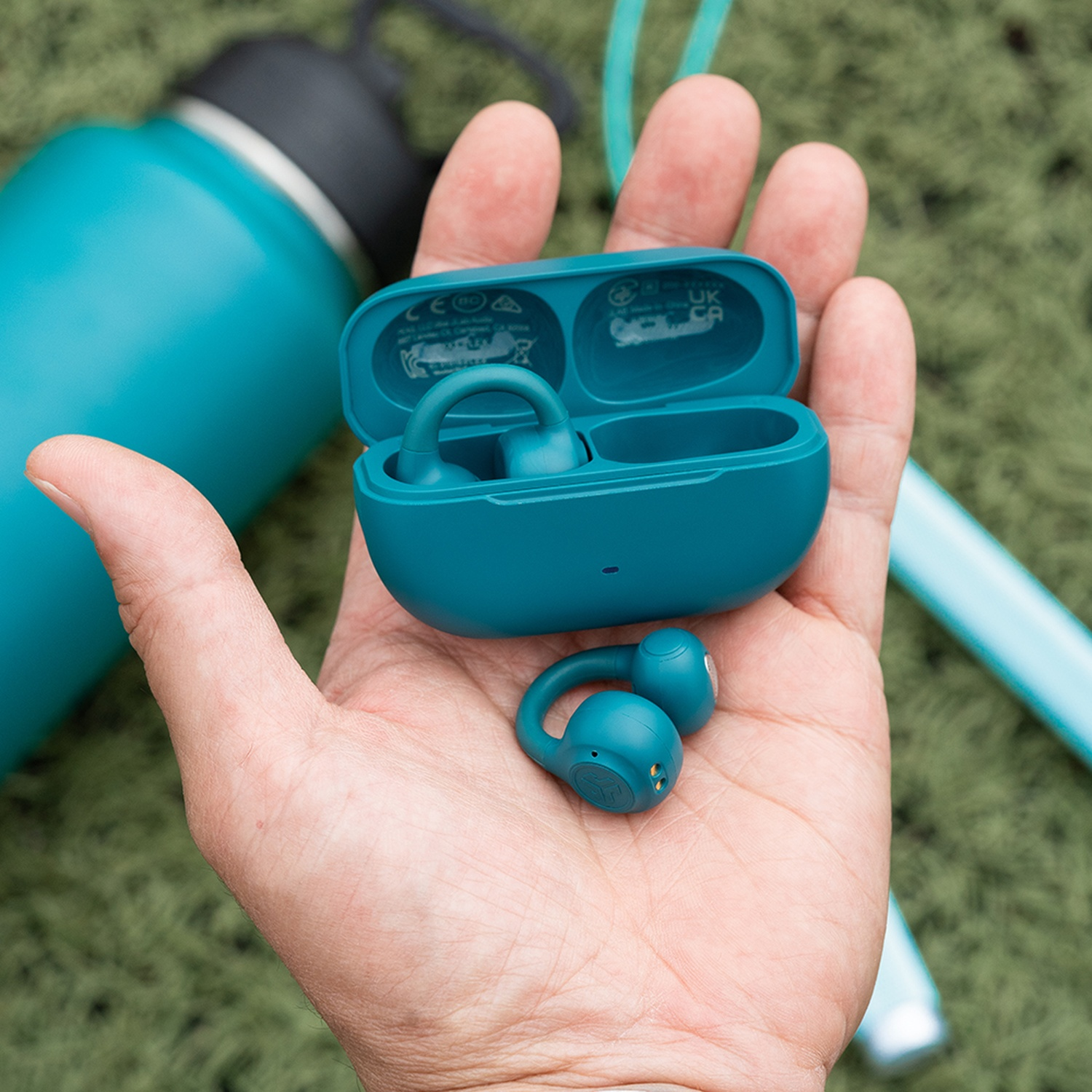 Une main tient des écouteurs sans fil et un étui turquoise. Une gourde turquoise et un tube turquoise sont en arrière-plan sur une surface verte.