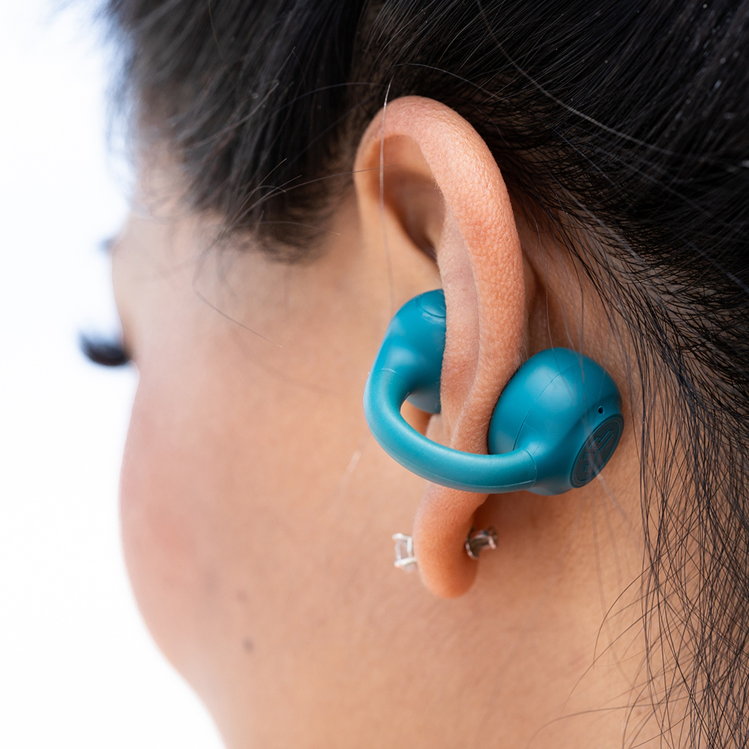 Une oreille avec un écouteur sarcelle. L'écouteur entoure l'oreille. On voit des cheveux foncés et de la peau.