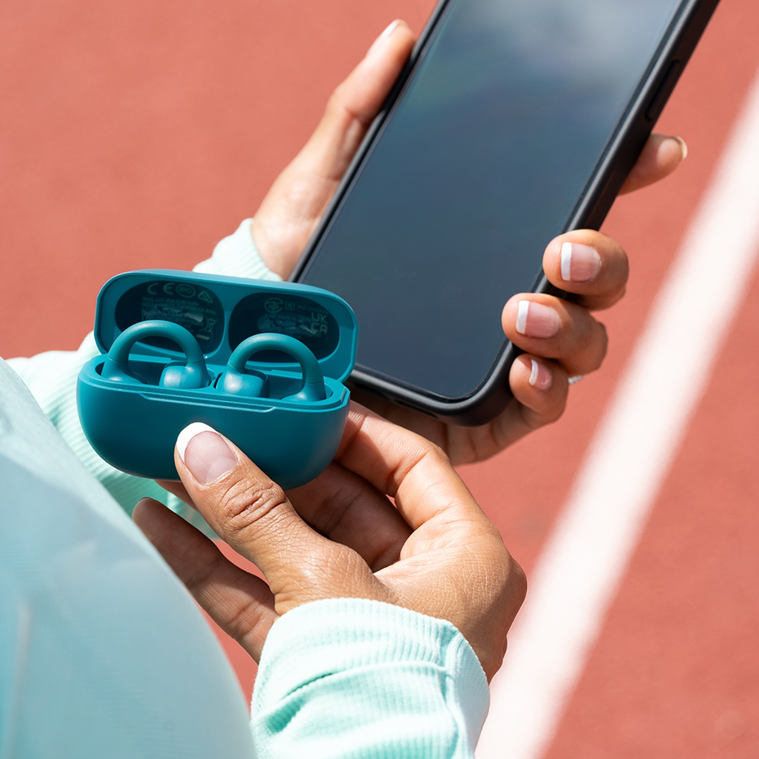 Une personne tient un téléphone et des écouteurs turquoise dans un étui. L'arrière-plan est une piste de course rouge. Les ongles ont des bouts blancs.