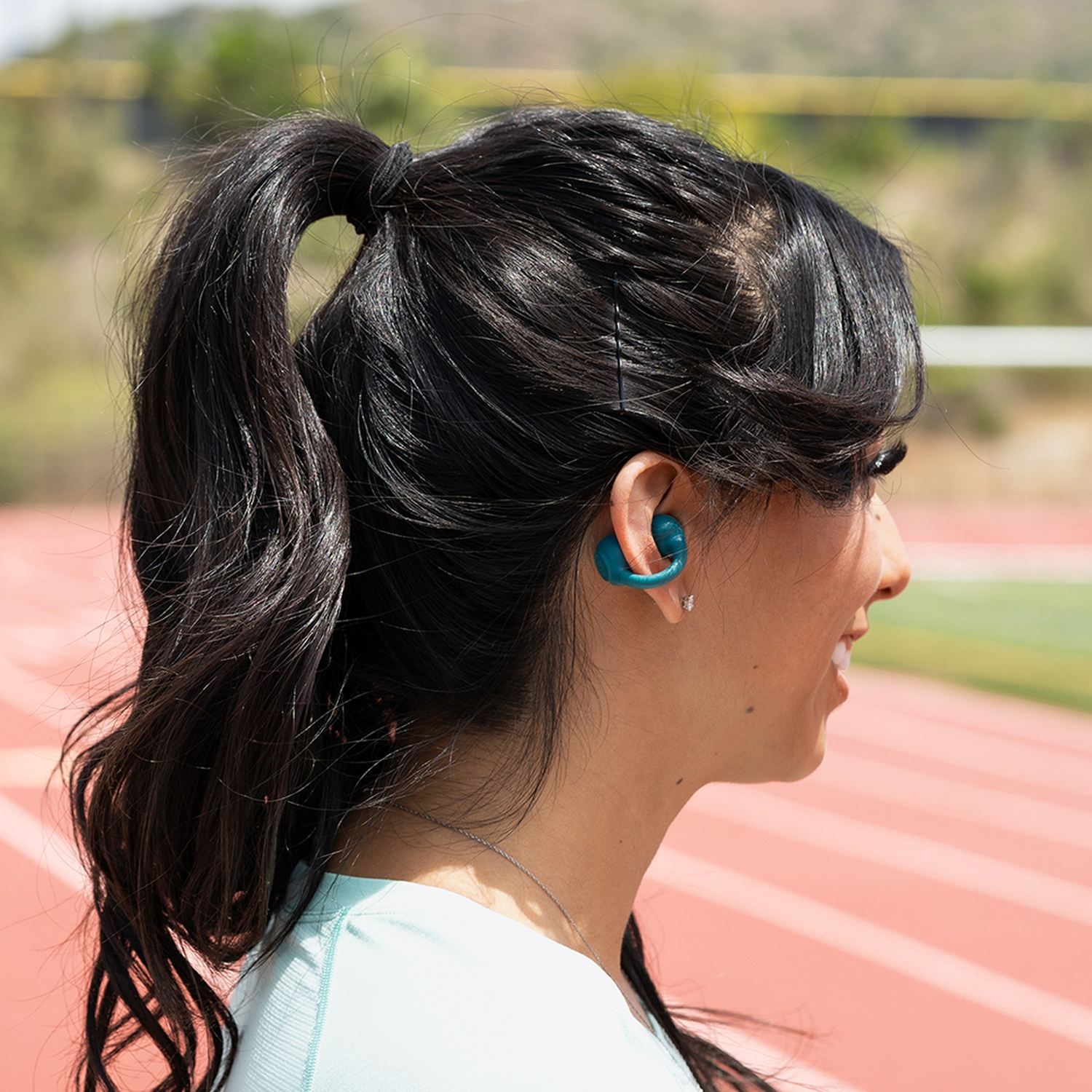Une femme aux cheveux noirs en queue de cheval porte des écouteurs turquoise. Elle sourit sur un terrain d'athlétisme. L'arrière-plan a des arbres verts.