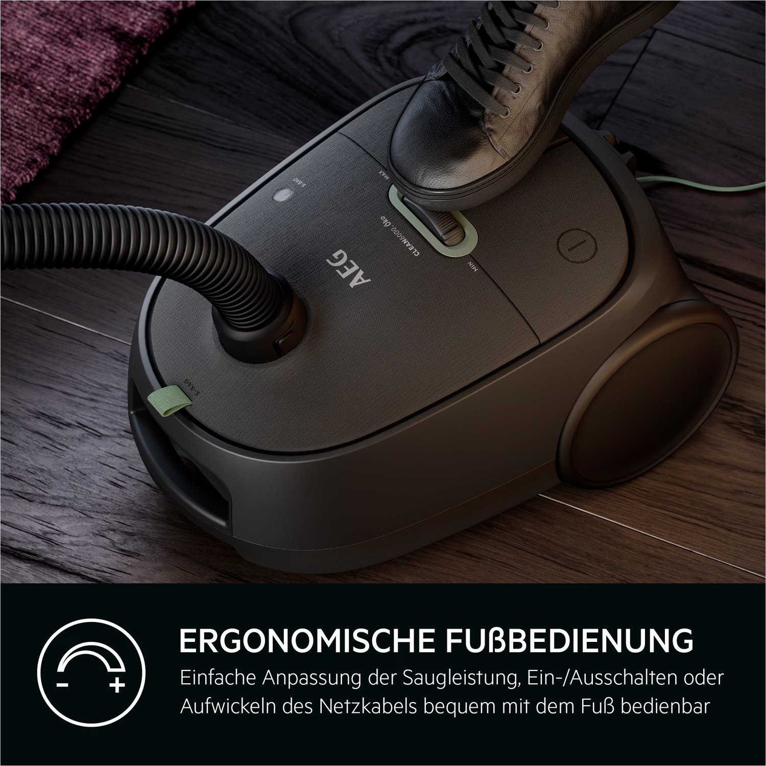 Black vacuum cleaner with a foot on top. It has a hose, and the brand is AEG. Wooden floor in the background.