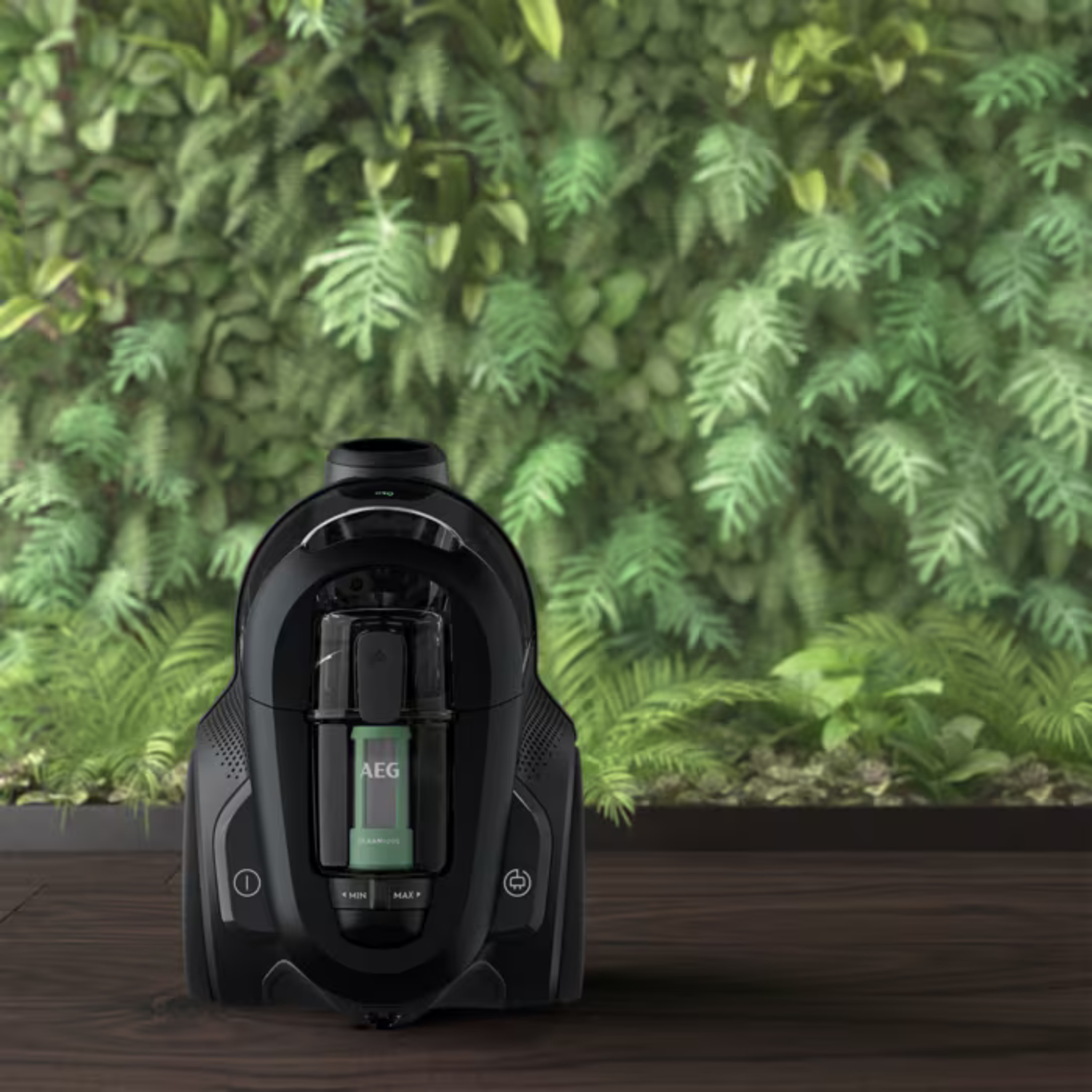 A black AEG vacuum cleaner is on a wooden floor. Behind it is a wall of green plants. The vacuum cleaner is turned on.