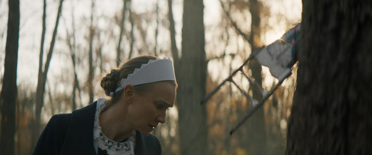 Eine Frau mit weißer Haube und schwarzem Mantel blickt in einem Wald nach unten. Im Hintergrund hängt eine Leiter. Bäume sind im Hintergrund.