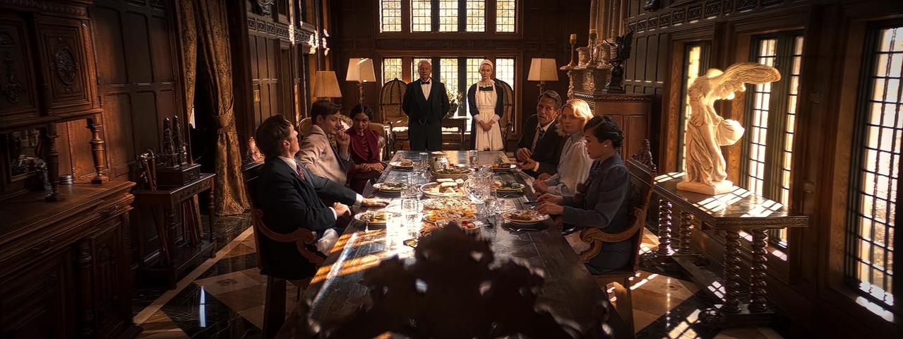 Eine Szene im Esszimmer. Menschen sitzen an einer langen Tafel für ein Essen. Im Hintergrund stehen zwei Kellner. Sonnenlicht scheint durch die Fenster.