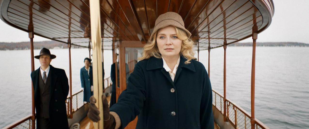 Eine Frau mit Hut und Mantel blickt auf einem Boot nach vorne. Zwei Männer stehen hinter ihr.