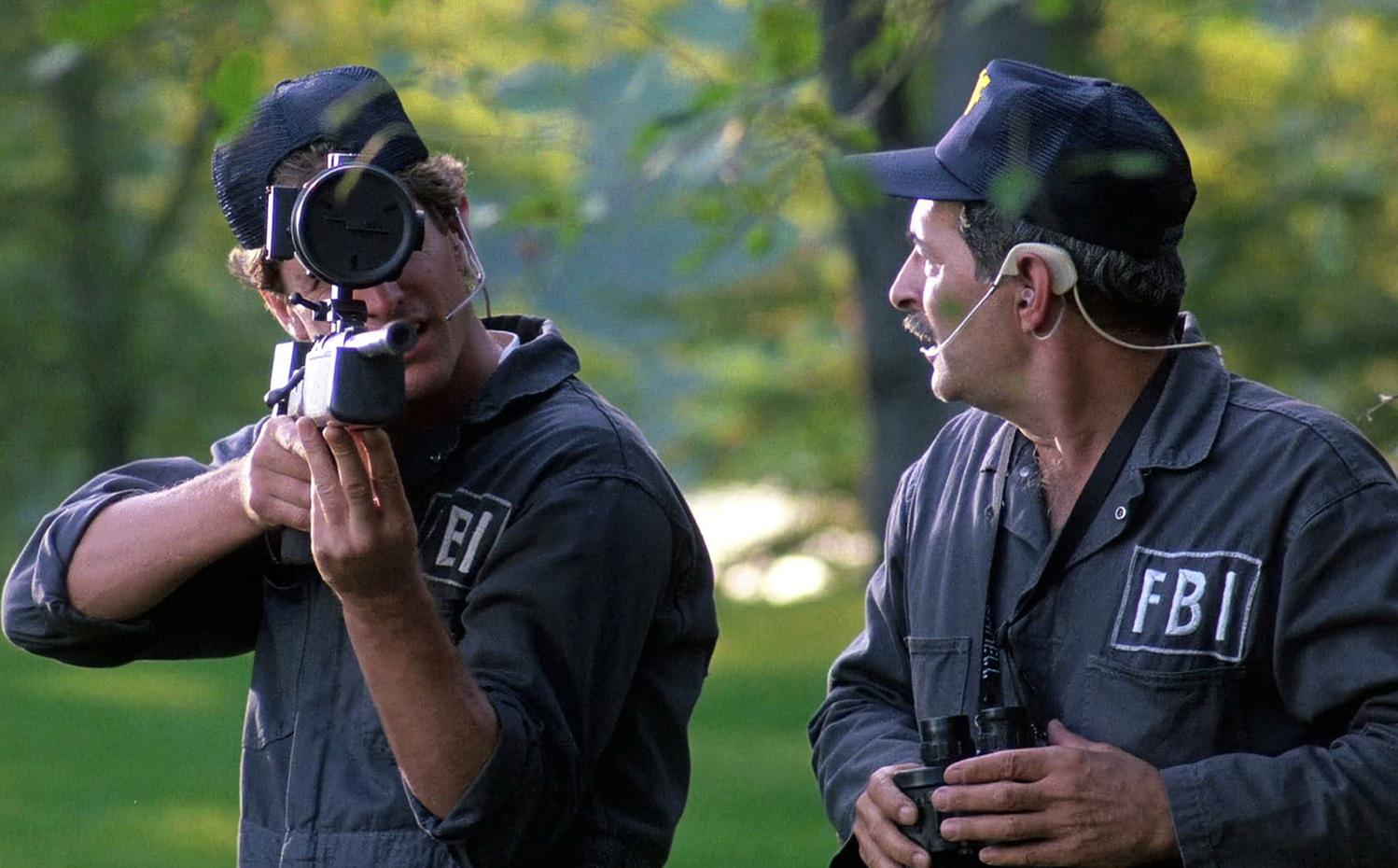Zwei Männer in FBI-Uniformen sind in einem Waldgebiet. Einer zielt mit einem Gerät, der andere hält ein Fernglas.