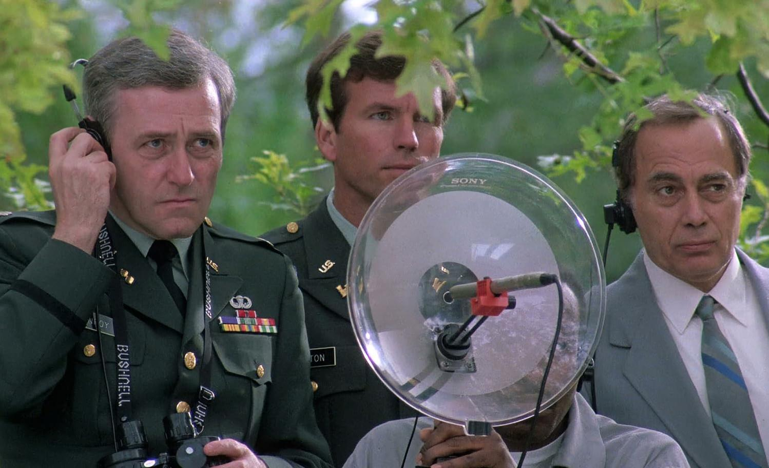 Drei Männer in Militäruniformen und Anzügen mit einem großen Parabolmikrofon. Einer benutzt ein Headset. Grünes Laub im Hintergrund.