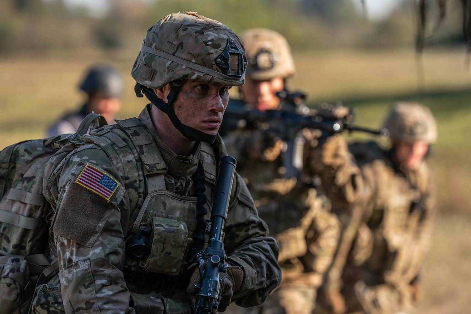 Soldat in Tarnung, Helm und Ausrüstung, hält ein Gewehr, amerikanischer Flaggenaufnäher. Andere Soldaten im Hintergrund.