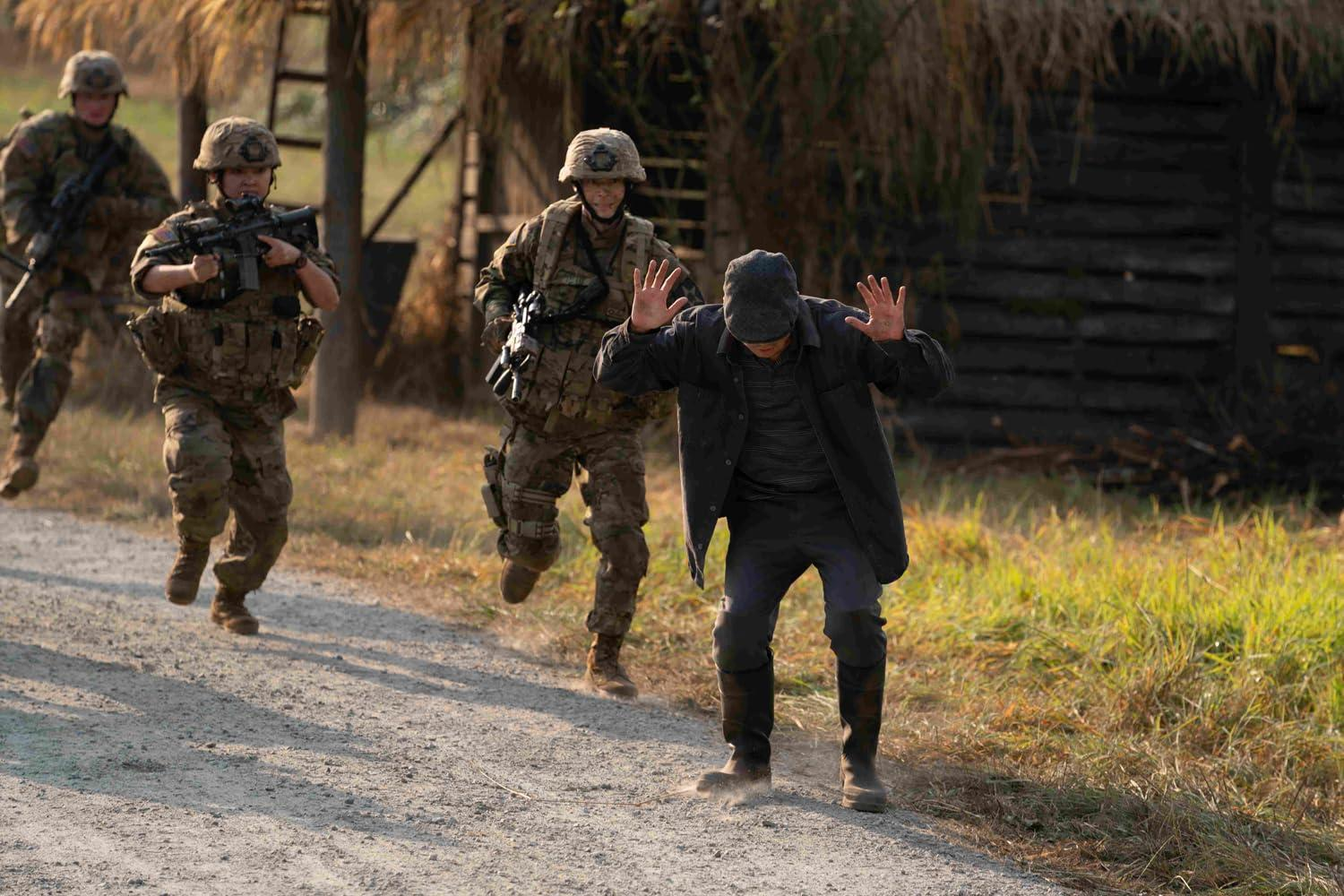 Soldaten mit Waffen verfolgen einen Mann mit erhobenen Händen. Der Mann trägt dunkle Kleidung. Soldaten tragen Helme und Tarnkleidung auf einer unbefestigten Straße.