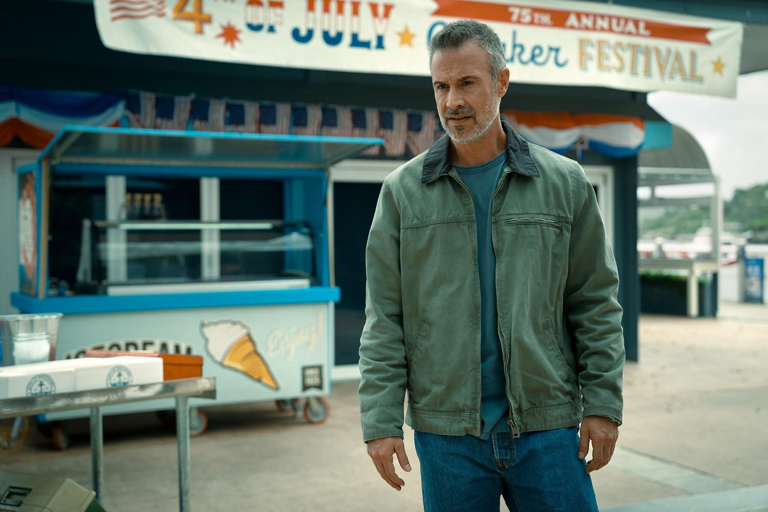 Ein Mann steht in der Nähe eines Eisstandes. Der Stand hat ein Banner zum 4. Juli. Der Mann trägt eine Jacke und Jeans.