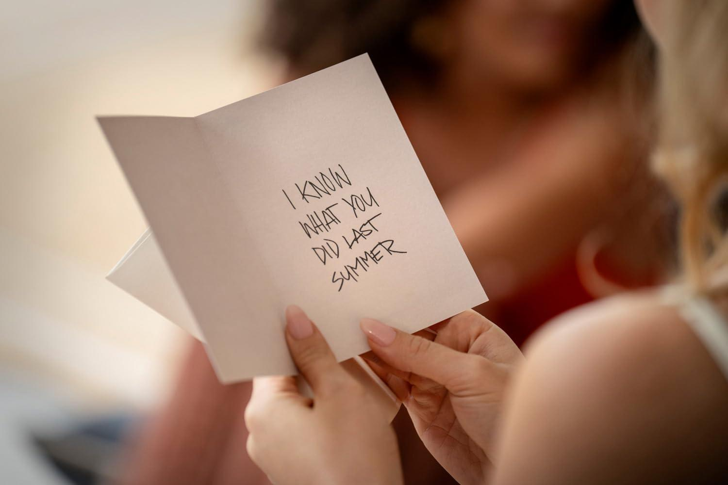 Eine Person hält eine Karte mit der Aufschrift „I KNOW WHAT YOU DID LAST SUMMER“. Andere Personen sind im Hintergrund verschwommen.