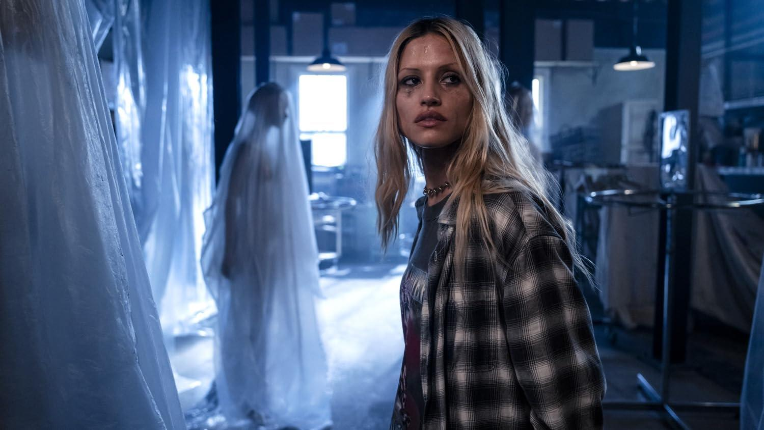 Eine Frau mit verschmiertem Make-up und einem karierten Hemd steht in einem dunklen Raum. Im Hintergrund eine mit weißem Stoff bedeckte Figur.