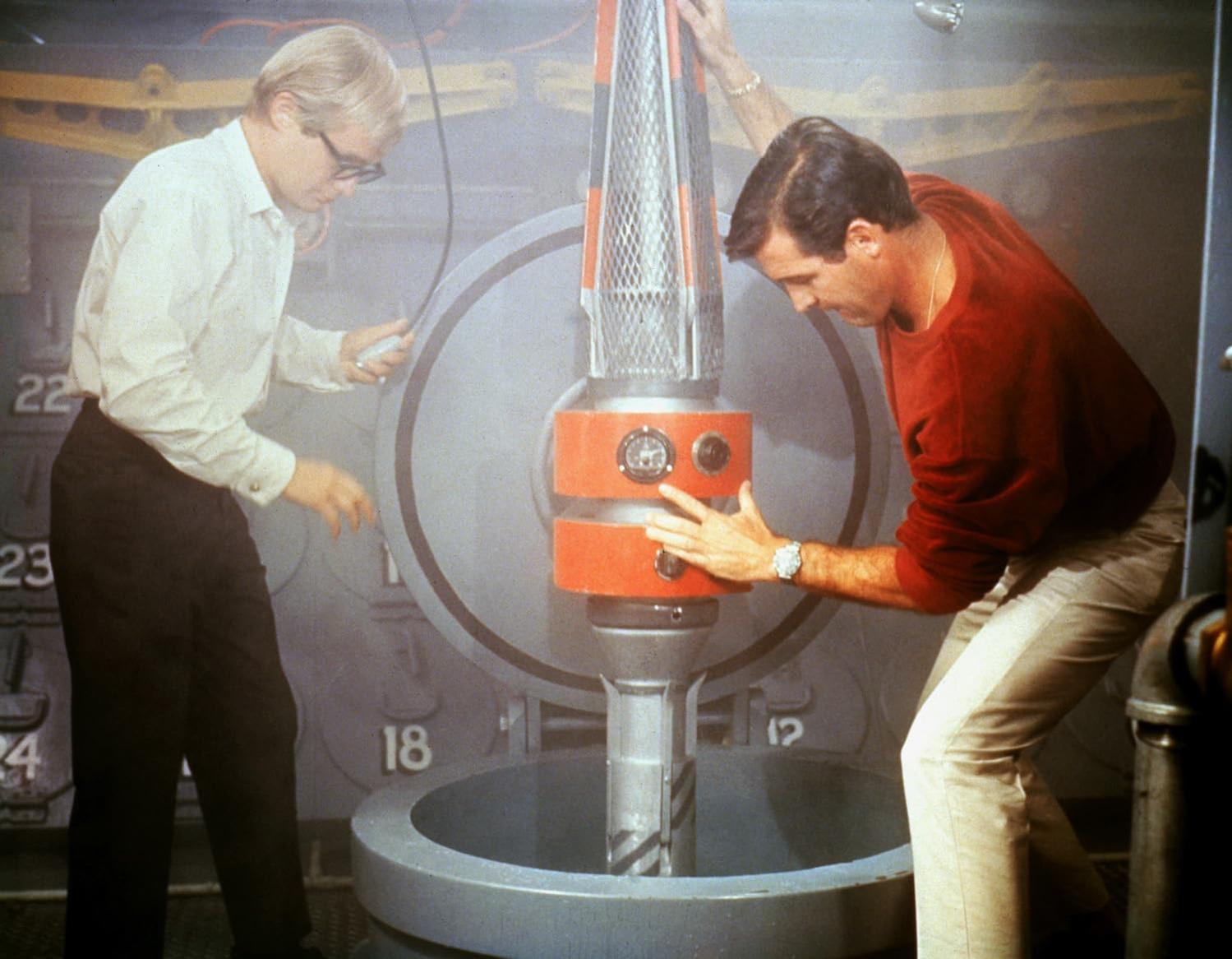 Zwei Männer untersuchen ein futuristisches Gerät. Einer trägt ein weißes Hemd, der andere einen roten Pullover. Das Gerät ist zylindrisch, mit orangefarbenen und silbernen Teilen.