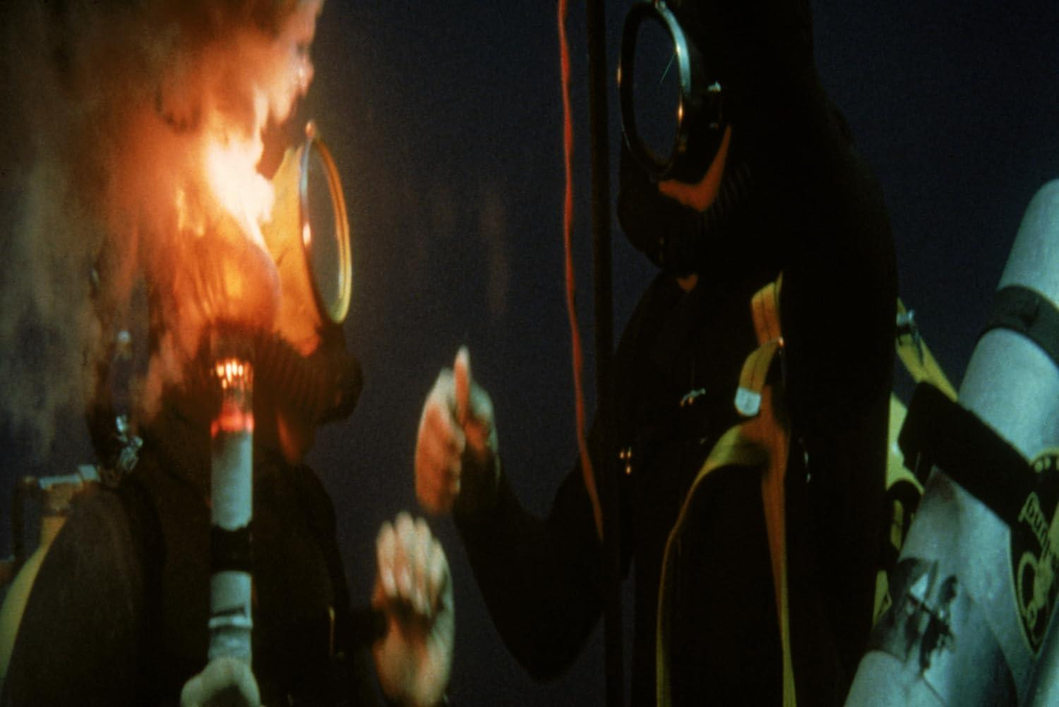 Taucher in dunklen Anzügen und Masken, einer hält eine brennende Fackel. Flammen und Rauch verdecken die Szene, Nachtkulisse.