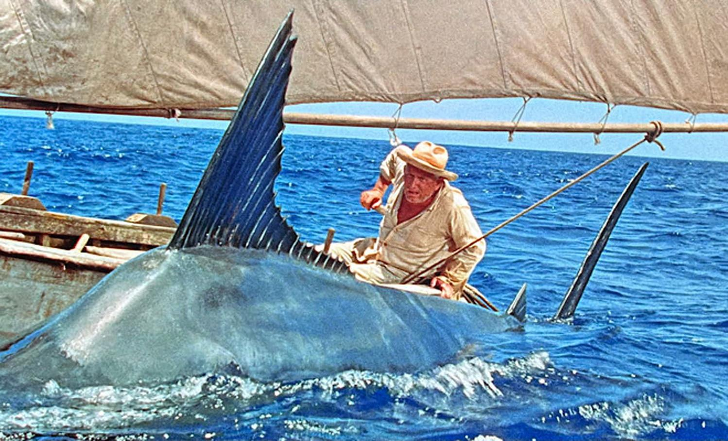 Ein älterer Mann kämpft in einem Boot mit einem großen Marlin. Die Flosse des Fisches ragt auf, das Segel ist sichtbar und das Meer ist blau.