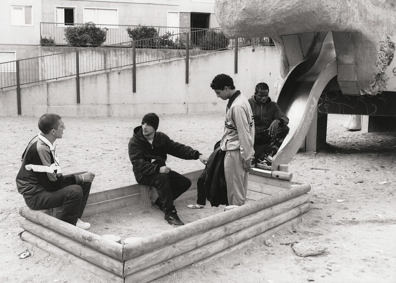 Vier junge Männer auf einem Spielplatz. Einer sitzt, zwei stehen an einer Rutsche, einer sitzt auf einer Holzkonstruktion. Schwarz-Weiß-Foto.
