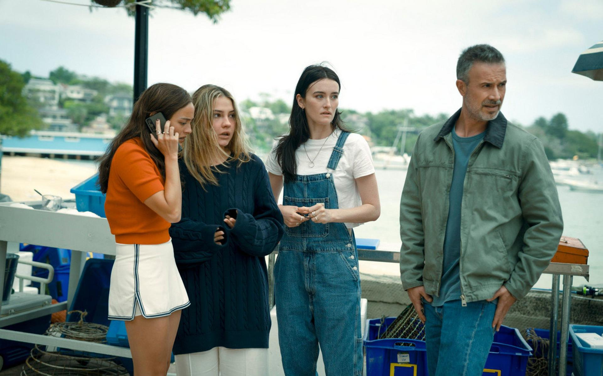 Vier Personen stehen in der Nähe des Wassers. Einer telefoniert. Andere sehen besorgt aus. Boote und Bäume sind im Hintergrund.