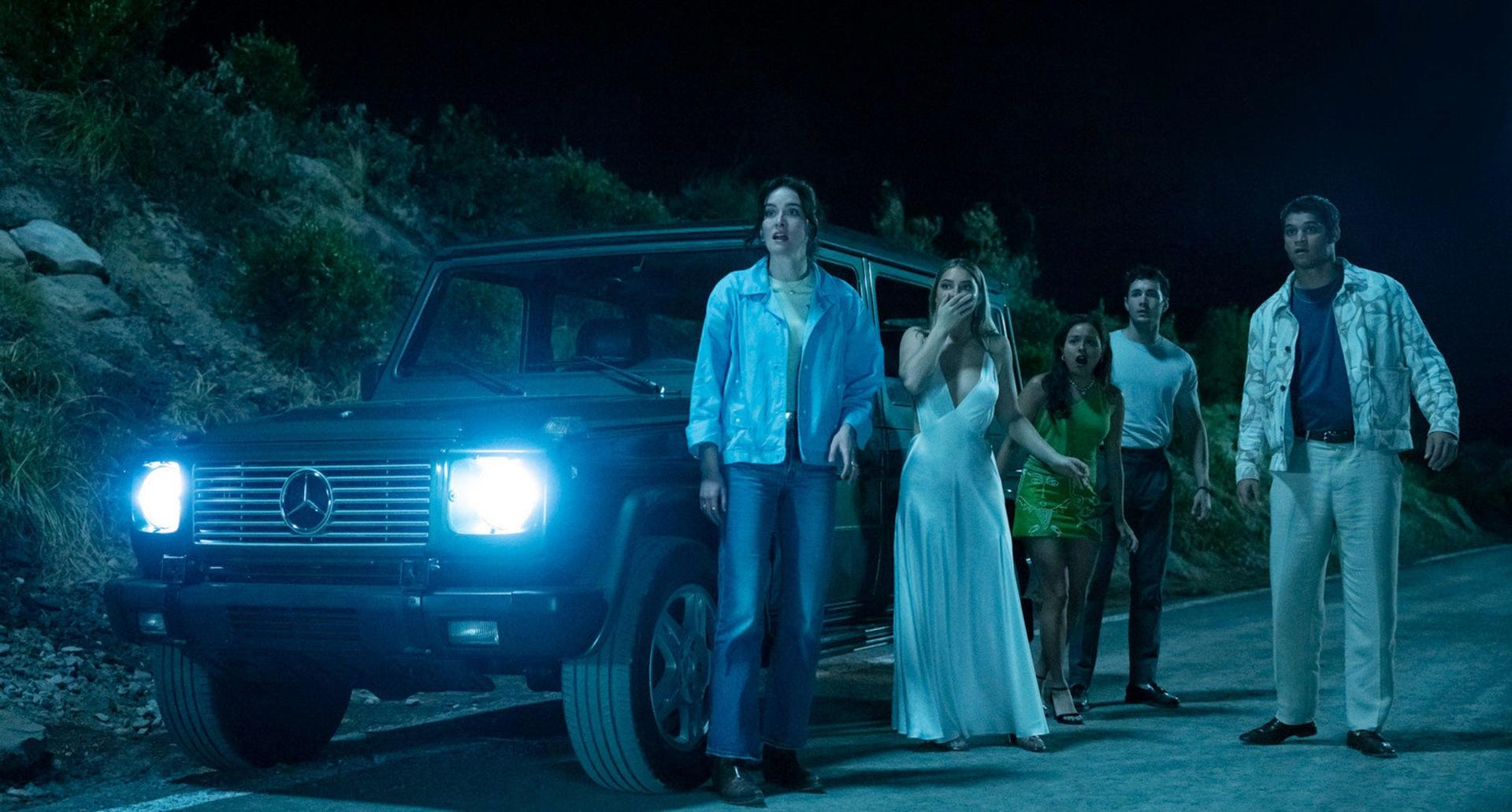 Gruppe von Menschen, die nachts von einem Auto schockiert sind. Vorne steht ein dunkler SUV mit hellen Scheinwerfern. Im Hintergrund eine dunkle, hügelige Straße.