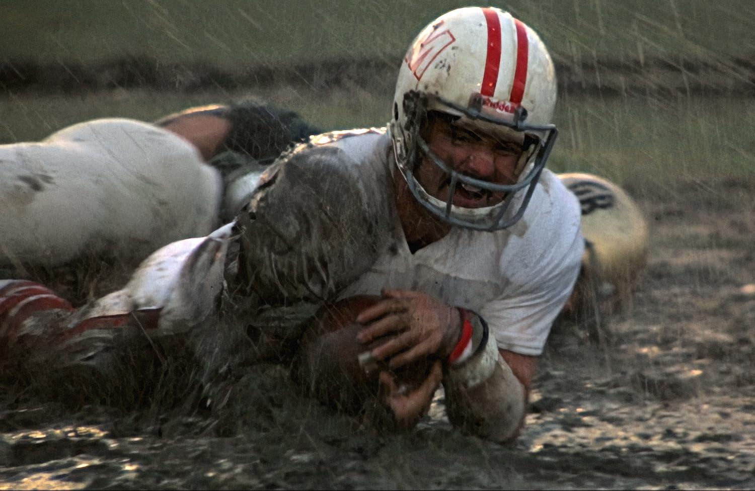 Ein Footballspieler, mit Helm, bedeckt mit Schlamm, umklammert den Ball am Boden. Andere Spieler und das Feld sind im Hintergrund.