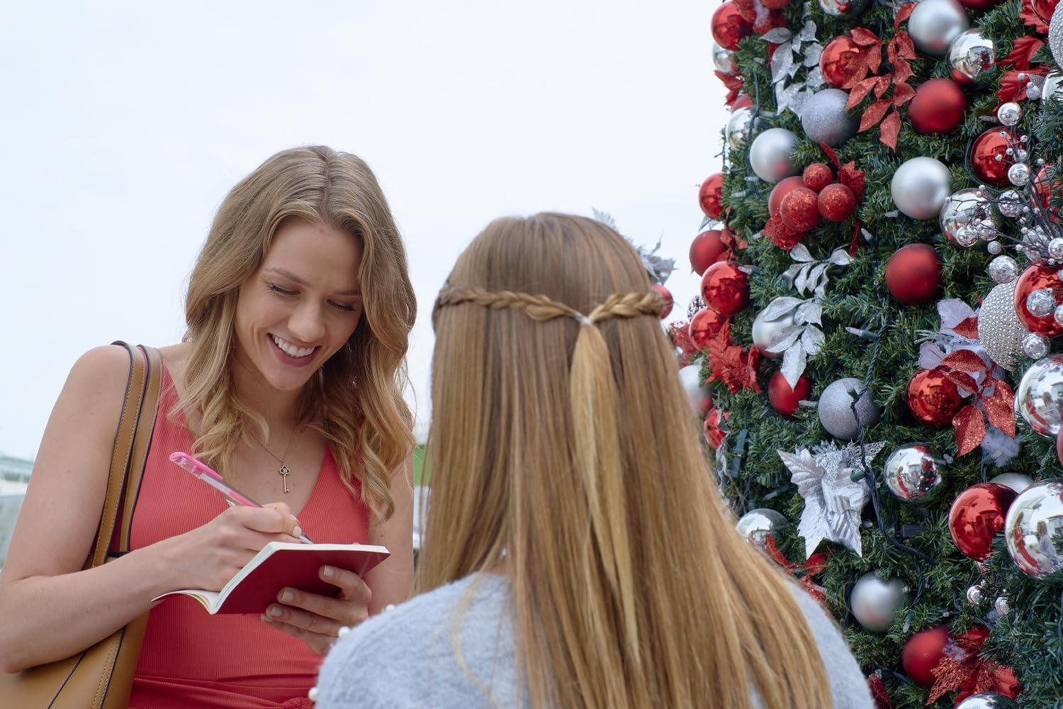 Zwei Frauen in der Nähe eines geschmückten Weihnachtsbaums. Eine schreibt lächelnd in ein Notizbuch. Der Baum hat rote und silberne Ornamente.