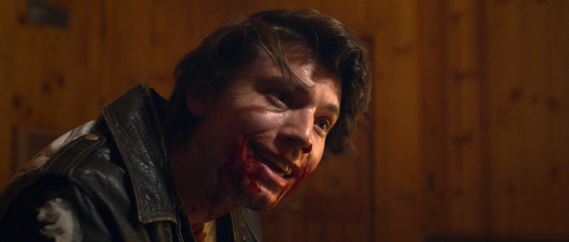 Ein Mann mit Blut um Mund und Gesicht lächelt. Er trägt eine Lederjacke und befindet sich in einem schwach beleuchteten Raum.