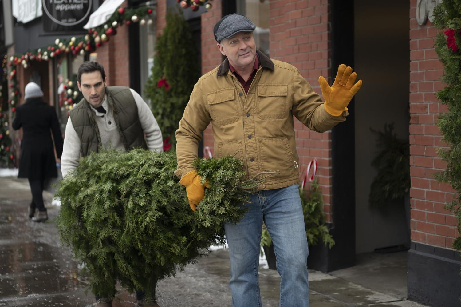 Zwei Männer tragen einen Weihnachtsbaum auf einer verschneiten Straße. Einer winkt, trägt eine braune Jacke und Mütze, der andere eine Weste.