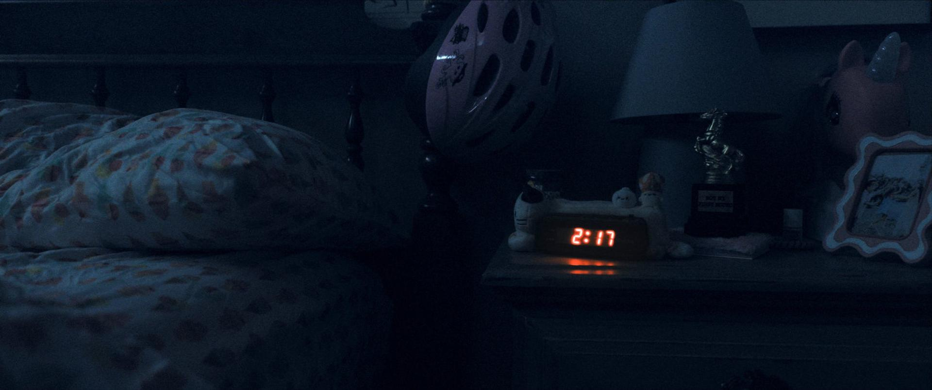 Ein schwach beleuchtetes Schlafzimmer bei Nacht. Bett mit gemusterten Bettlaken. Nachttisch mit Wecker, der 2:17 anzeigt, Lampe und kleinen Spielzeugen.