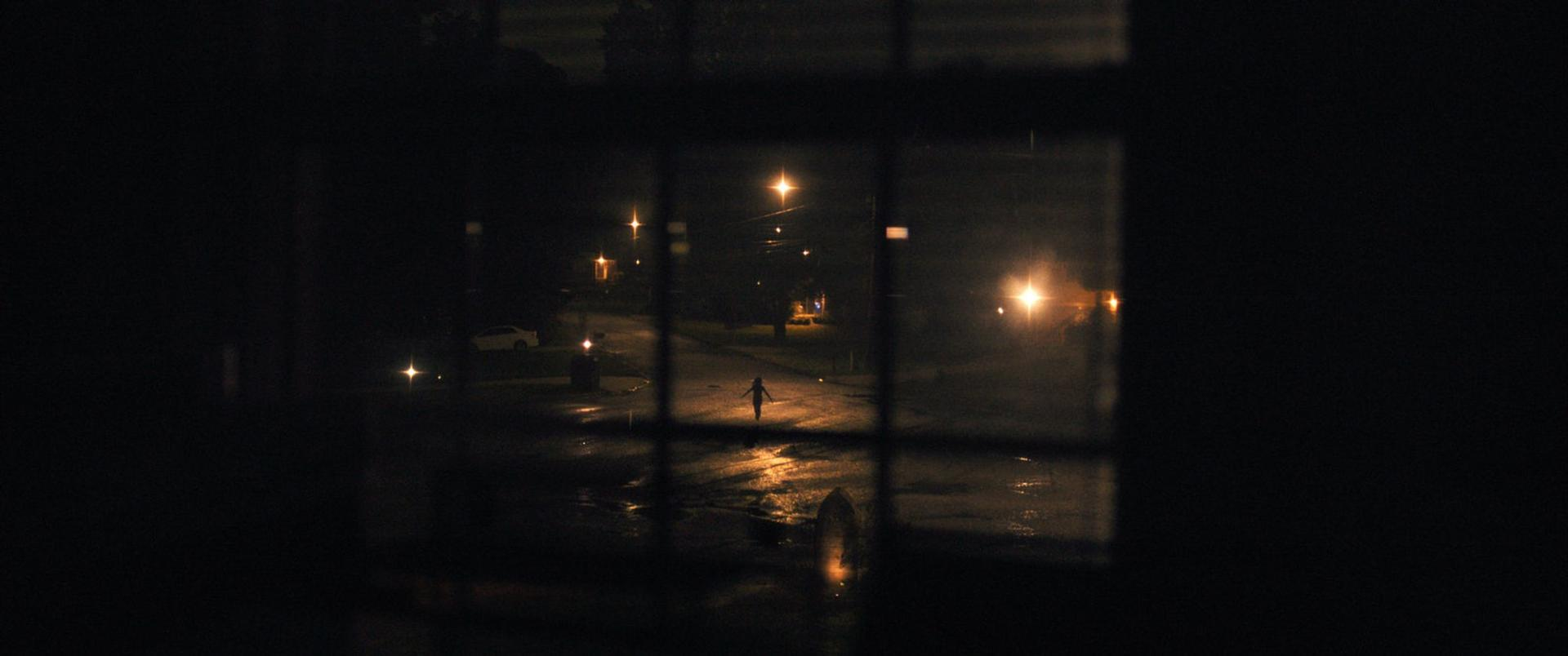 Nachtszenen durch ein Fenster. Eine Figur geht auf einer nassen Straße mit Straßenlaternen. Ein Auto ist geparkt.