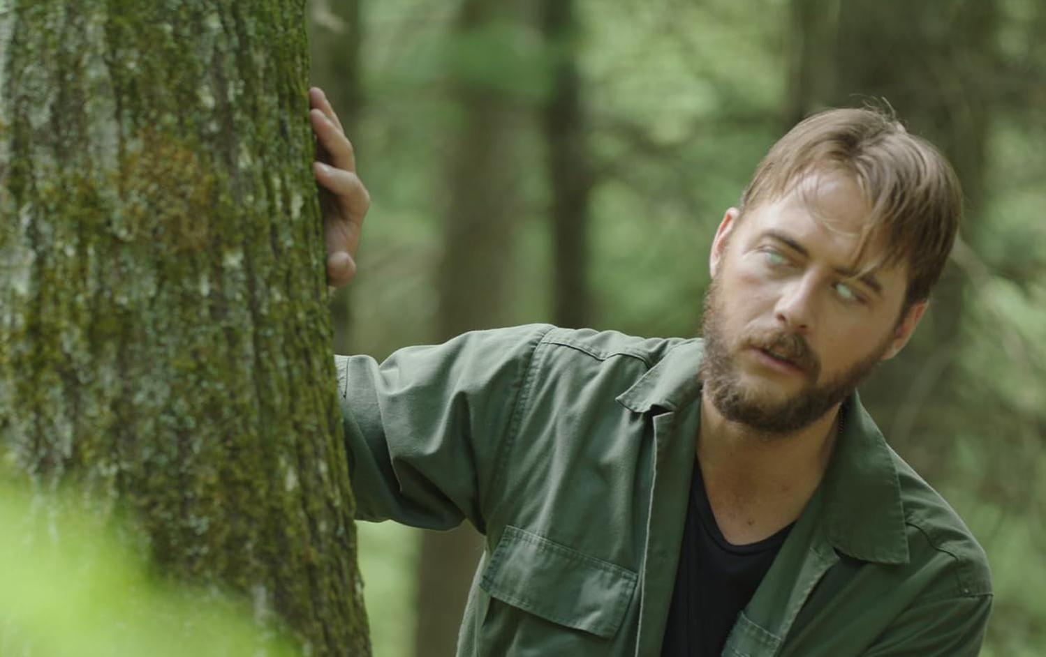 Ein Mann mit Bart, in einer grünen Jacke, schaut nachdenklich, während er sich an einen Baum lehnt.