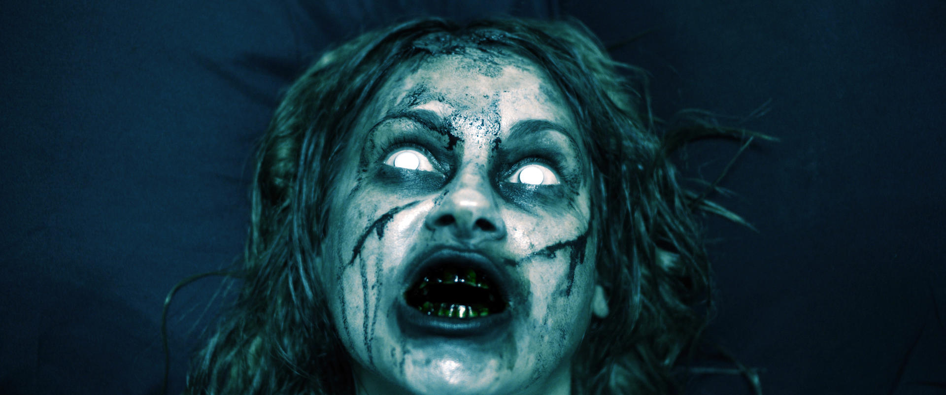 Ein Zombiegesicht mit leuchtend weißen Augen, offenem Mund mit grüner Substanz und dunklen Streifen auf der Haut vor dunklem Hintergrund.