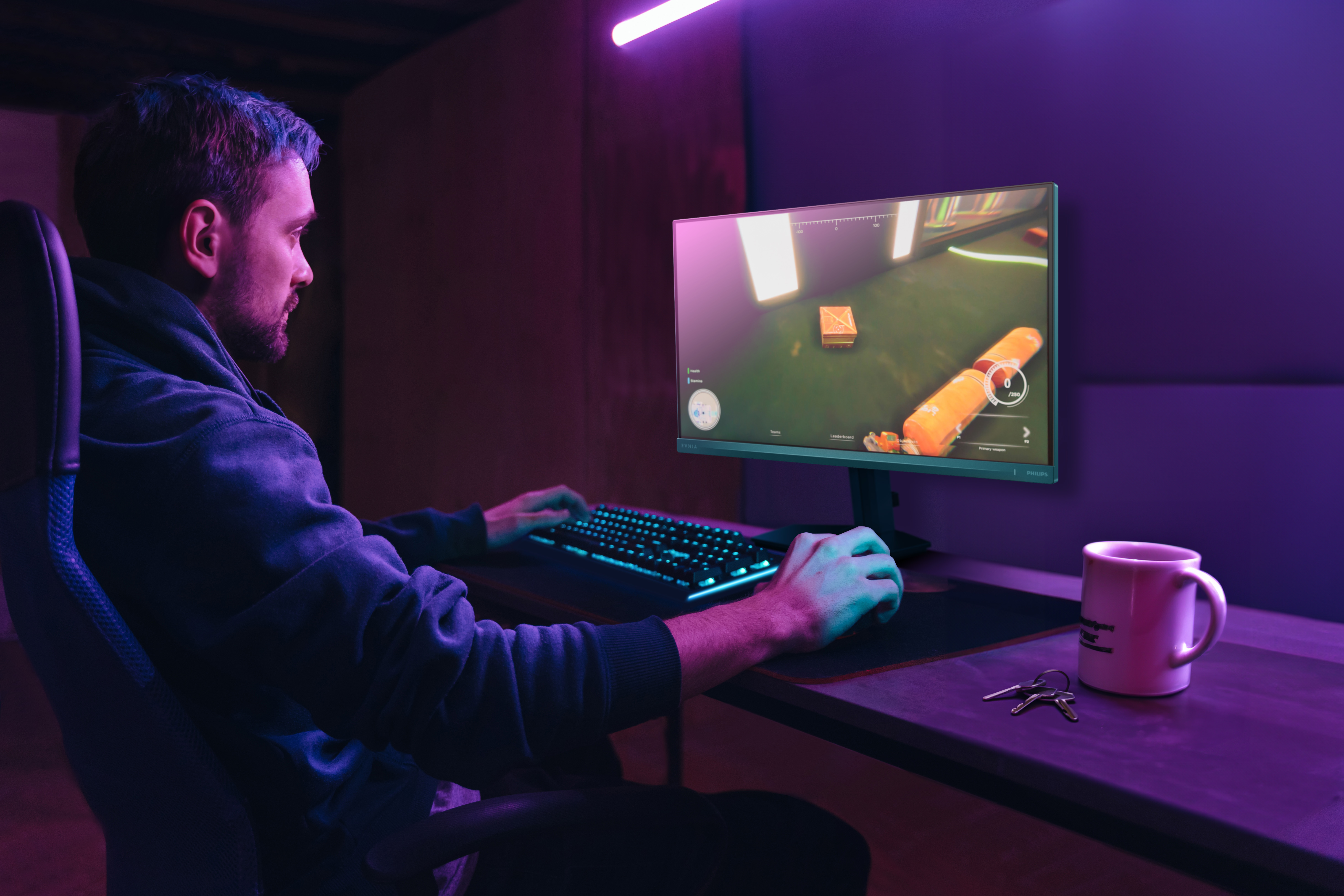 Eine Person spielt in einem dunklen Raum am Computer. Ein Monitor zeigt ein Spiel. Auf dem Schreibtisch stehen eine Tasse und Schlüssel.