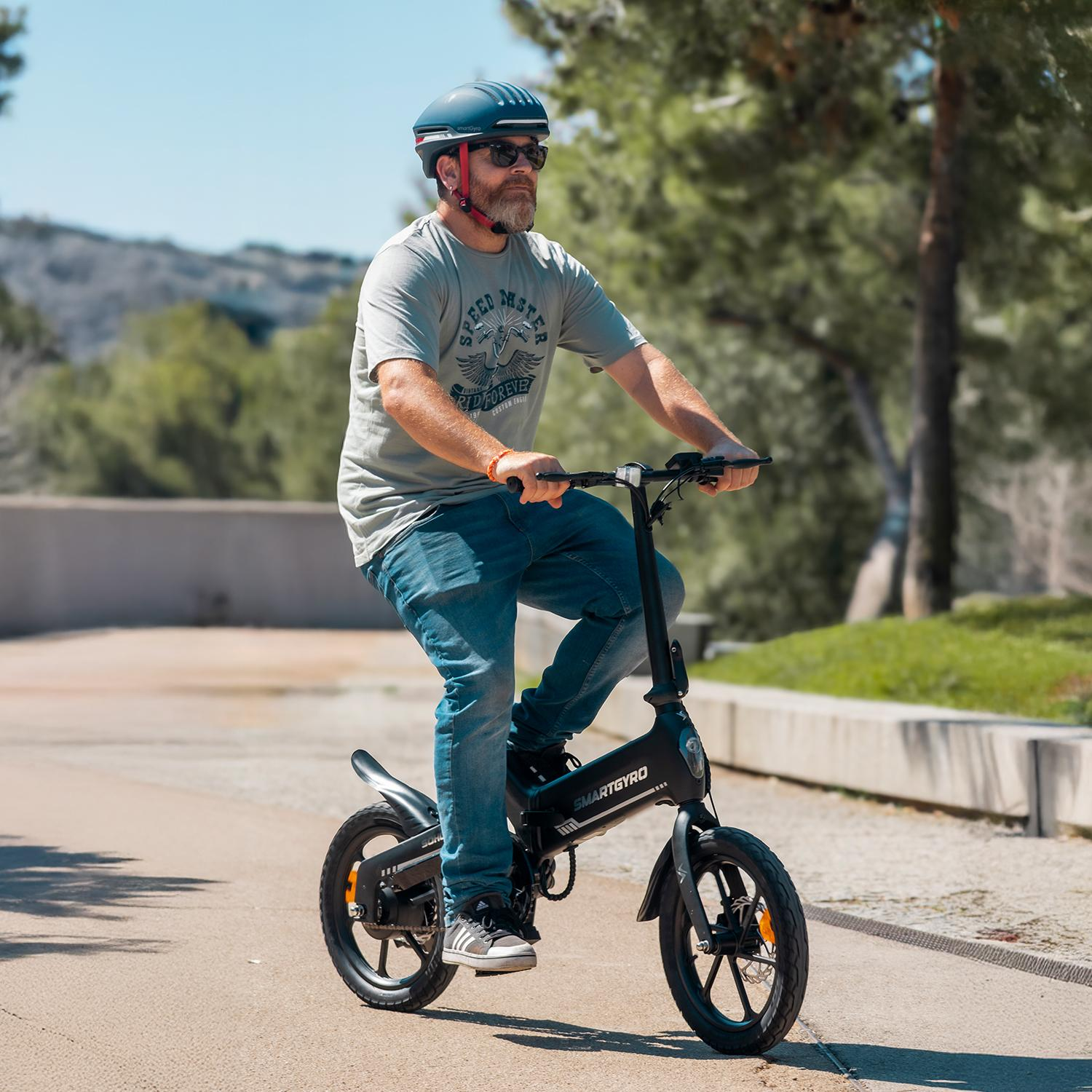 Un hombre monta una e-bike Smartgyro negra en un camino. Lleva casco, gafas de sol y vaqueros. Hay árboles y cielo en el fondo.