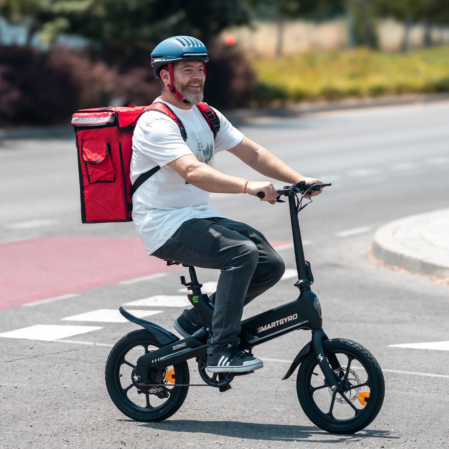 Un hombre monta en una bicicleta eléctrica negra. Lleva casco, una bolsa de reparto roja y ropa oscura. La bicicleta está en la carretera.