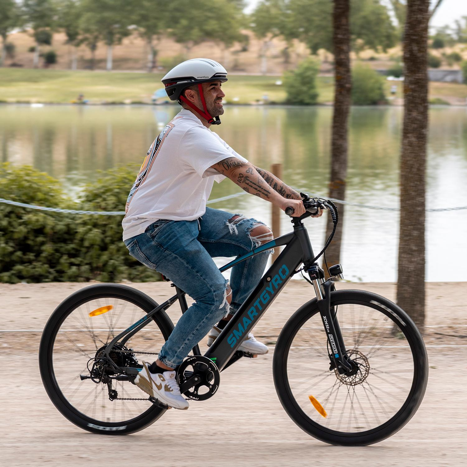 Un hombre monta en una bicicleta Smartgyro negra por un camino cerca de un lago. Lleva casco, camisa blanca y vaqueros. Árboles y agua al fondo.