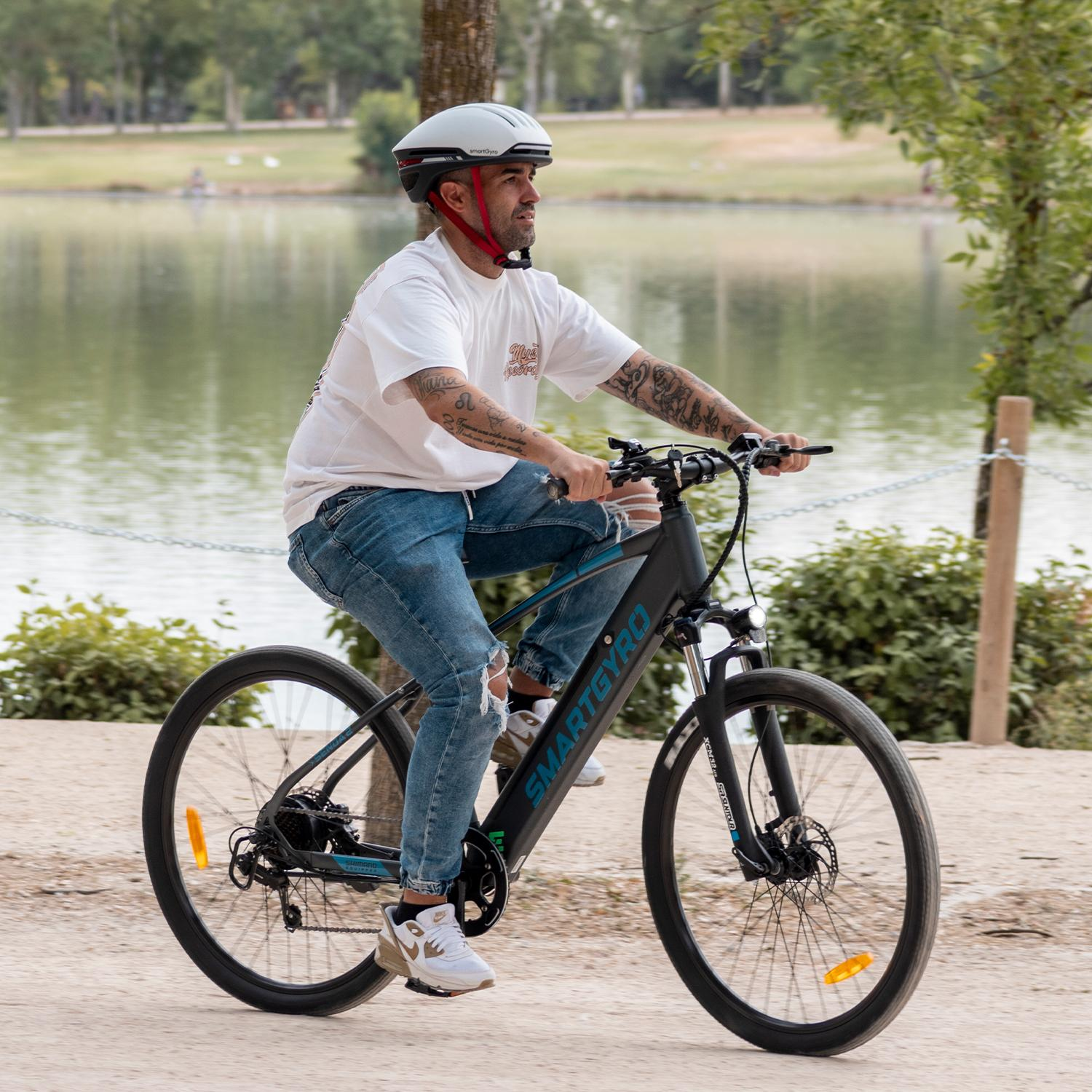 Hombre monta bicicleta Smartgyro negra cerca de un lago, usando casco y jeans.