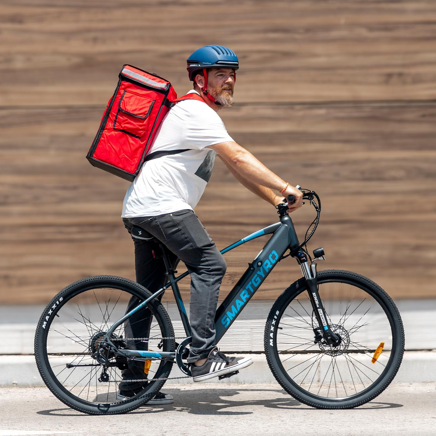 Un hombre monta en una bicicleta eléctrica con una bolsa de reparto roja. Lleva un casco azul y una camisa blanca. El fondo es una pared de madera.
