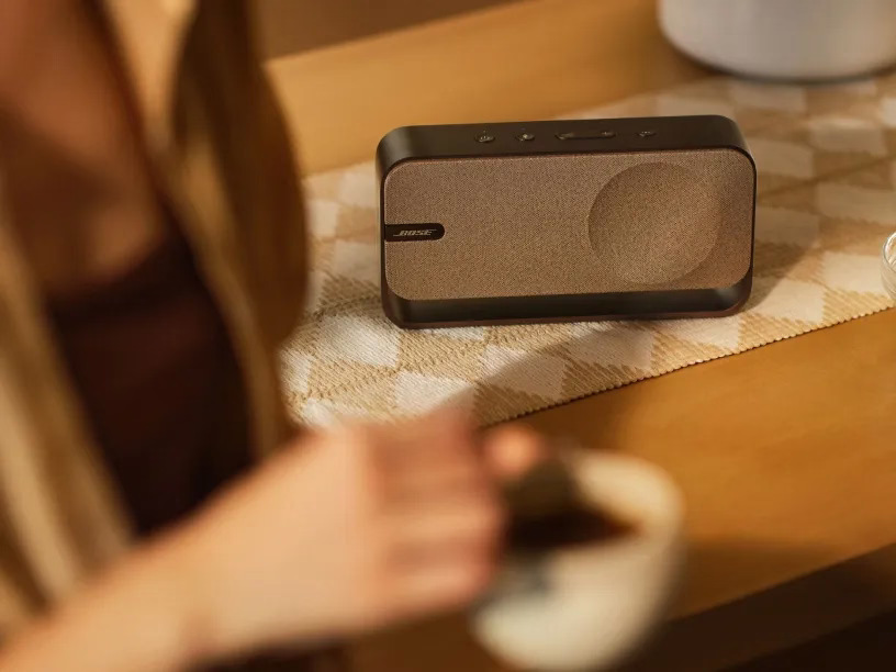 Brązowy i czarny głośnik stoi na stole. Osoba trzyma filiżankę kawy, tło jest rozmyte.