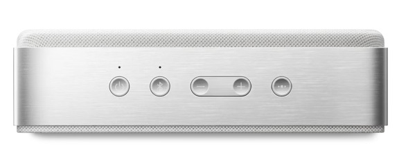 Srebrny głośnik z materiałowym wierzchem. Ma przyciski: zasilanie, Bluetooth, regulacja głośności i wyciszenie. Tło jest białe.