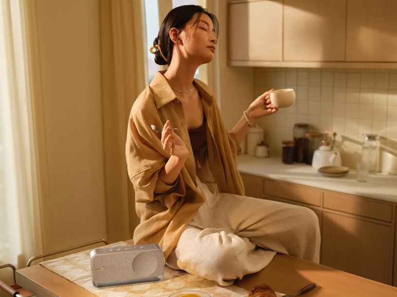 Kobieta siedzi z zamkniętymi oczami, trzymając filiżankę. Głośnik na stole. Kuchnia w tle. Miękkie światło.
