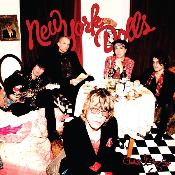 A New York Dolls együttes látható. Öt tag egy szobában van egy asztallal, italokkal és egy sakktáblás padlóval.