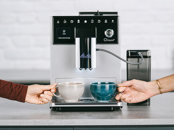 İki elin fincan tuttuğu bir kahve makinesi. Bir el bej bir fincanı, diğeri ise turkuaz bir fincanı tutuyor. Makine gümüştür.