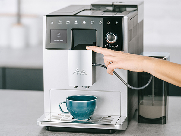 Bir kişinin eli kahve makinesine dokunuyor. Altında mavi bir kupa var. Sağda süt kabı. Makine gümüş ve siyah.