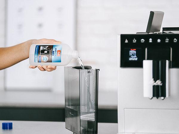 Bir kişi kahve makinesinin su deposuna kireç çözücü sıvı döküyor. Makine beyaz ve siyahtır.