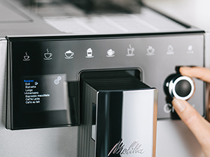 Bir kahve makinesinin kontrol panelinin yakın çekimi, bir el kadranı çeviriyor. Simgeler ve bir ekran görülebilir.