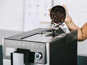 Bir kişi kahve çekirdeklerini bir kahve makinesine döküyor. Makine gri ve siyah. Arka plan bulanık.